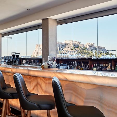 Electra Metropolis Hotel Athena Bagian luar foto The photo features a modern bar area with a sleek design. It showcases a long bar counter made of a light-colored material, possibly a type of stone, that has a smooth, polished finish. Behind the bar, there are shelves lined with various bottles, su