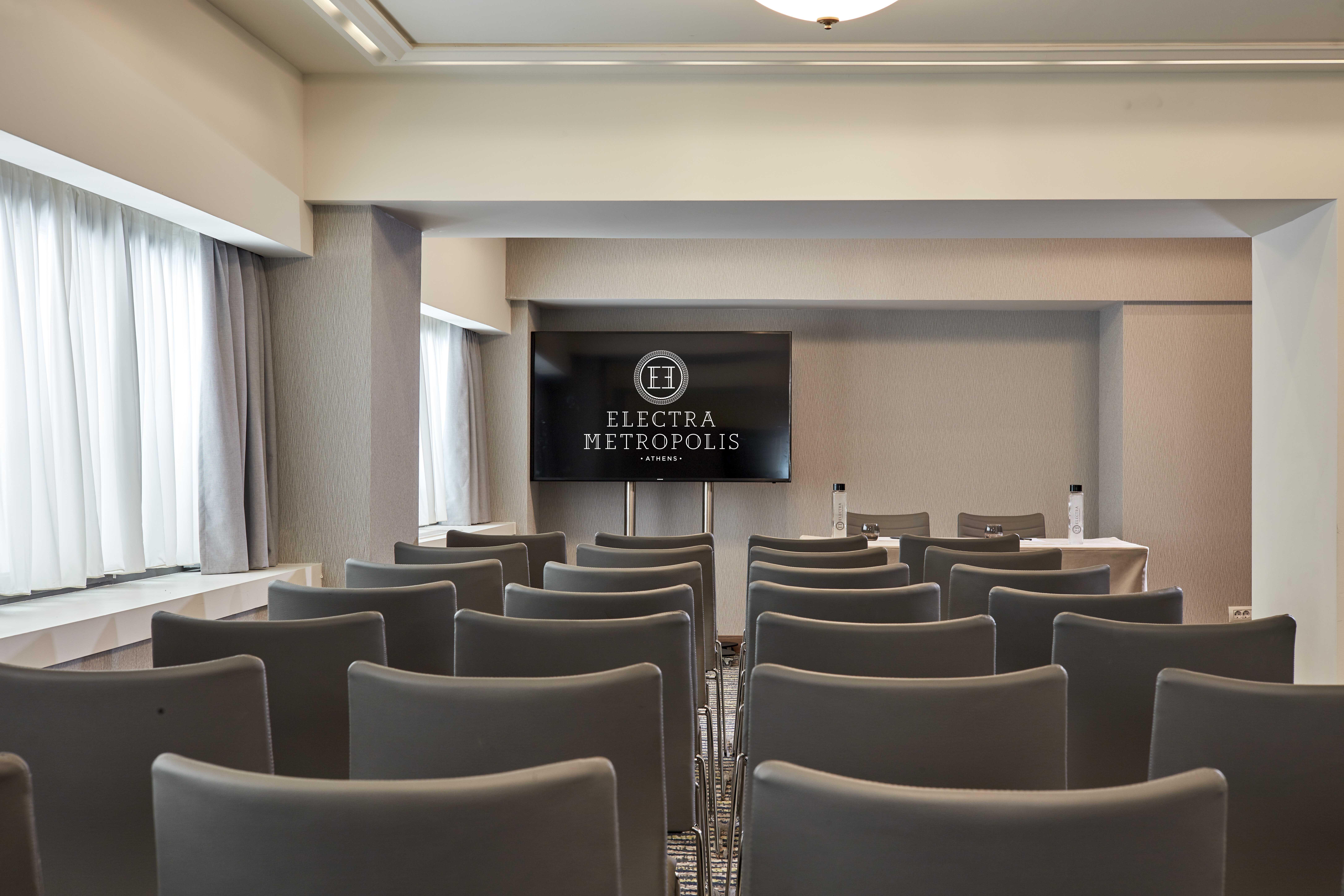 Electra Metropolis Hotel Athena Bagian luar foto The photo shows a meeting or conference room. There are several rows of gray chairs arranged facing a large screen displaying the logo "ELECTRA METROPOLIS." The room has large windows that allow natural light in, and the walls are decorated in neutra