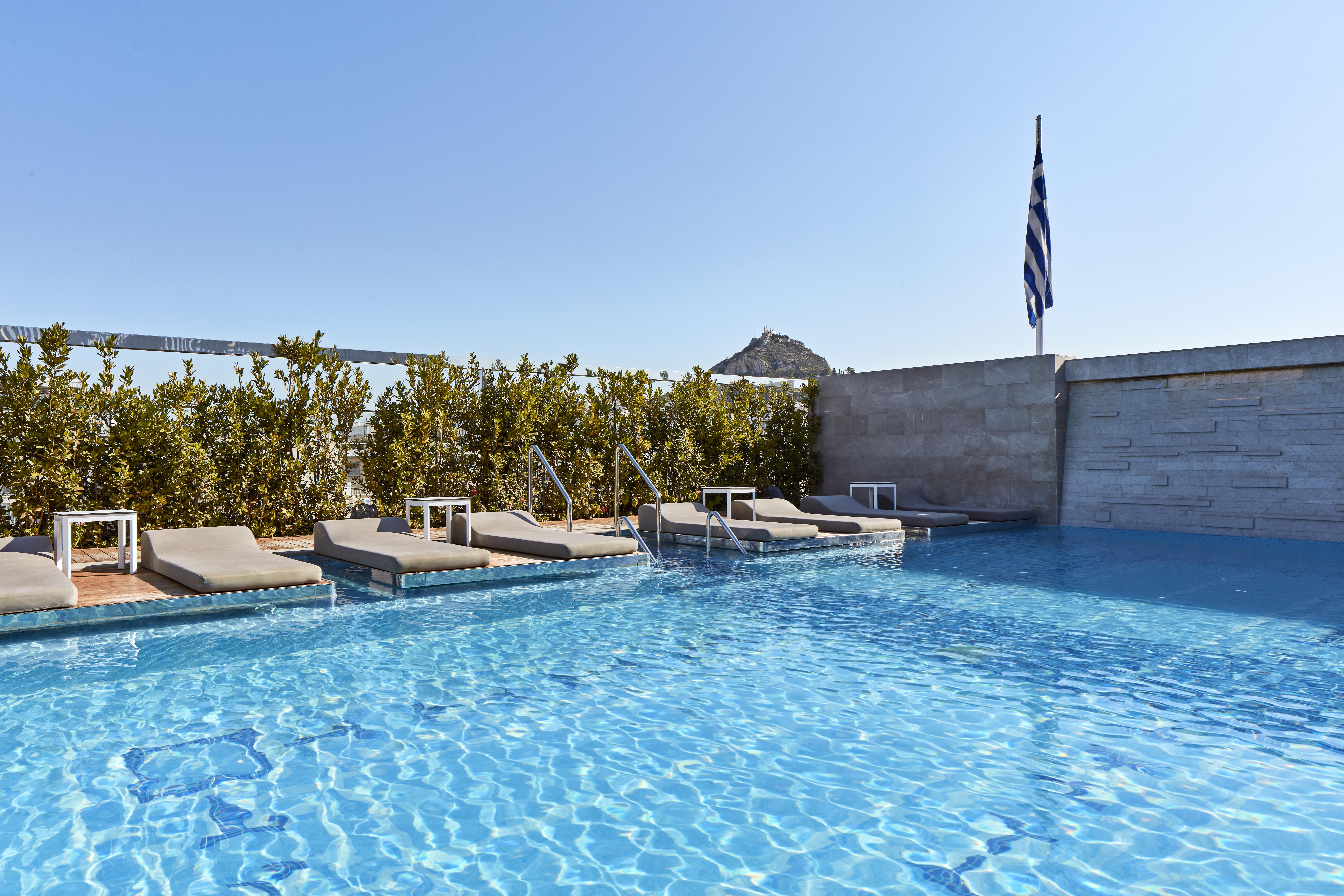 Electra Metropolis Hotel Athena Bagian luar foto The photo shows a rooftop swimming pool area. The water in the pool is clear and inviting, reflecting the blue sky above. There are several lounge chairs positioned around the pool, suggesting a relaxing atmosphere. In the background, there are green