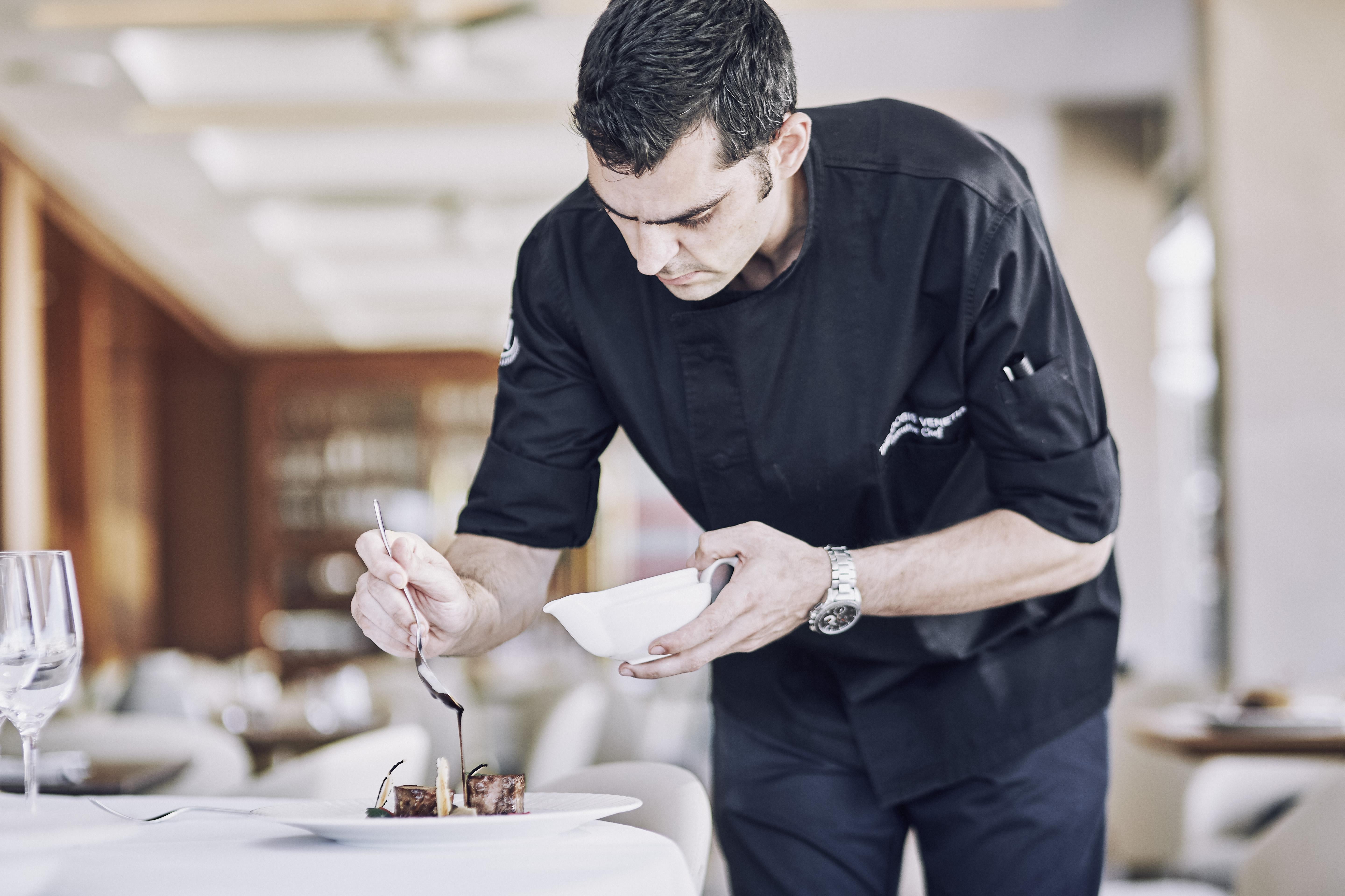 Electra Metropolis Hotel Athena Bagian luar foto The photo shows a chef in a black chef's jacket carefully plating a dish. He is leaning over a table, focused on adding a finishing touch to the presentation with a spoon. The dining area has a modern and elegant ambiance, with a soft focus on the ba