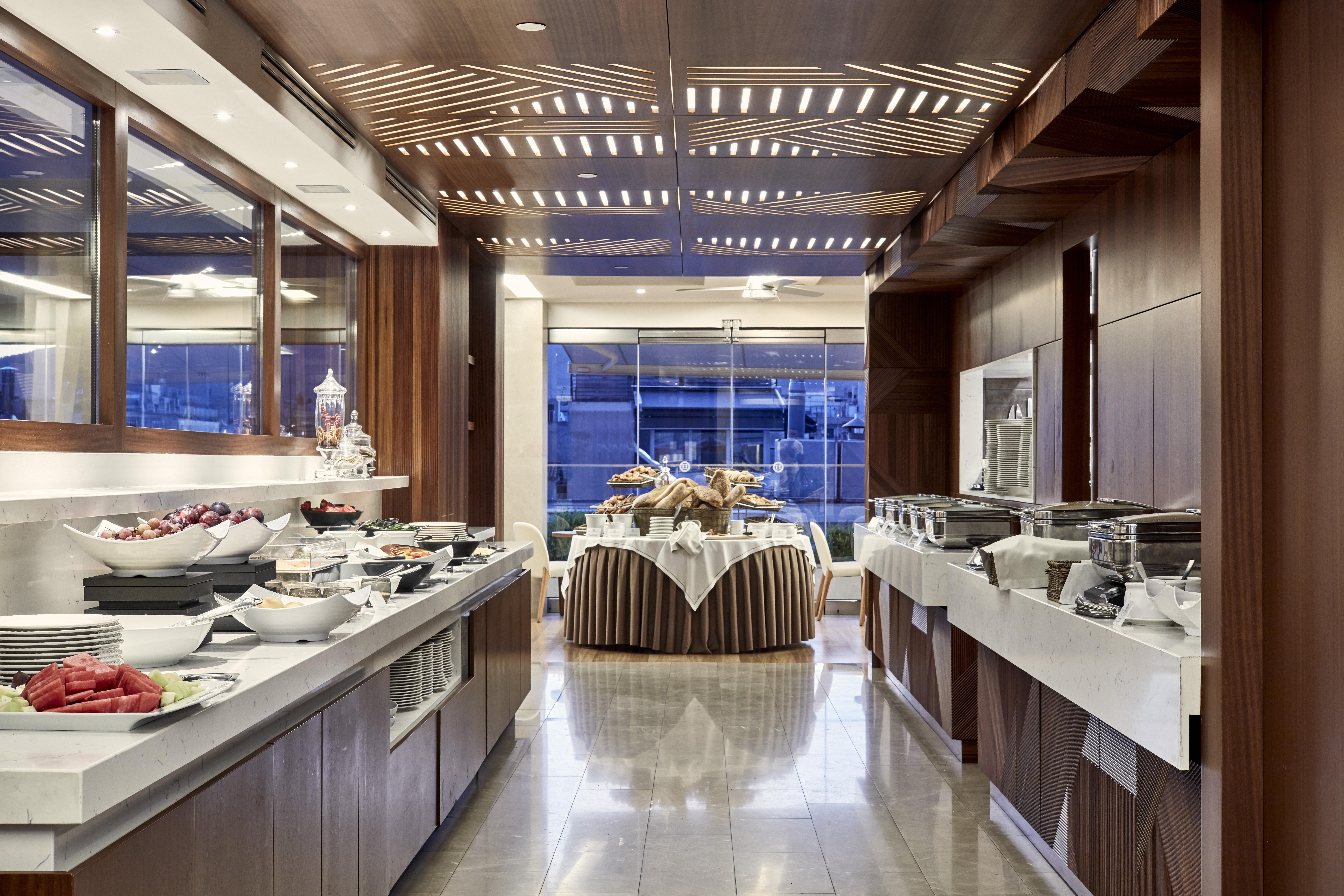 Electra Metropolis Hotel Athena Bagian luar foto The photo shows the interior of a modern dining area or buffet setup. The space features elegant wooden paneling and a contemporary design. On one side, there are several buffet tables with an assortment of food items, including fruits and salads, ne