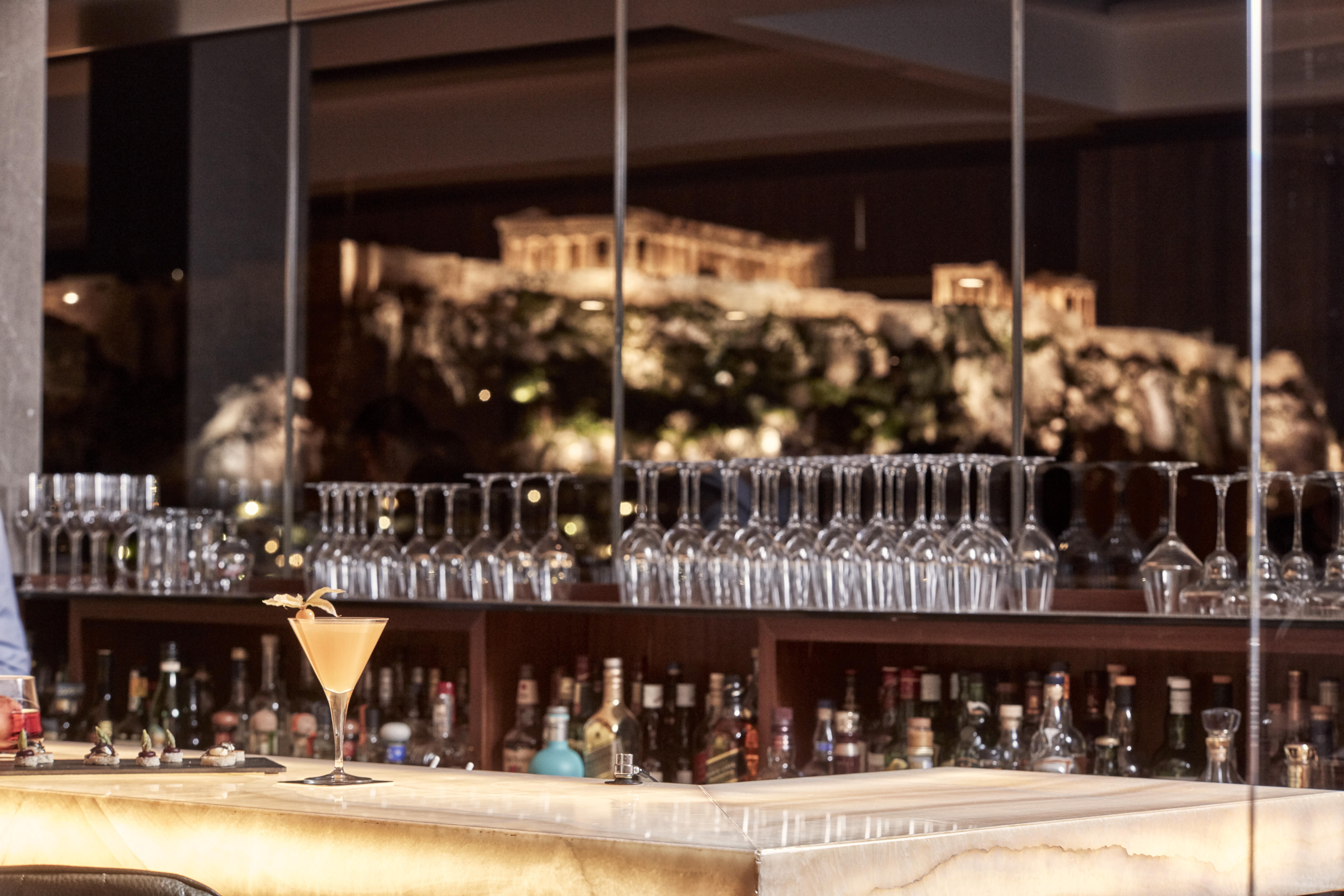 Electra Metropolis Hotel Athena Bagian luar foto The photo showcases a sophisticated bar setup, featuring a marble counter with a cocktail placed elegantly on it. Behind the bar, there are numerous glassware items neatly arranged on shelves, alongside various bottles of alcohol. In the background, 