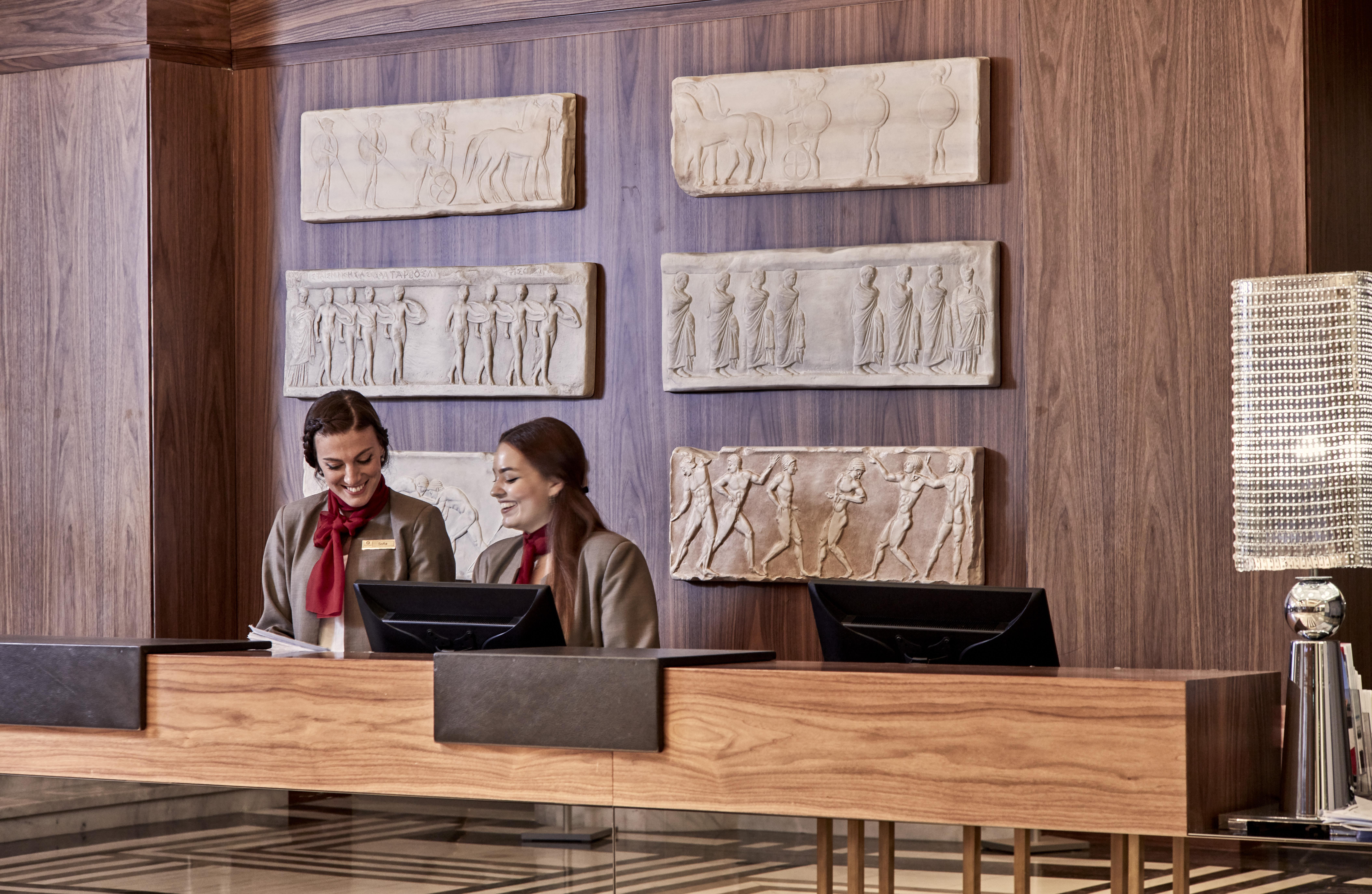 Electra Metropolis Hotel Athena Bagian luar foto The photo depicts a hotel reception area featuring two women at the front desk. They are engaged in conversation or checking something on a device. The reception desk is made of wood and has a modern design. Behind them, there are several relief scul
