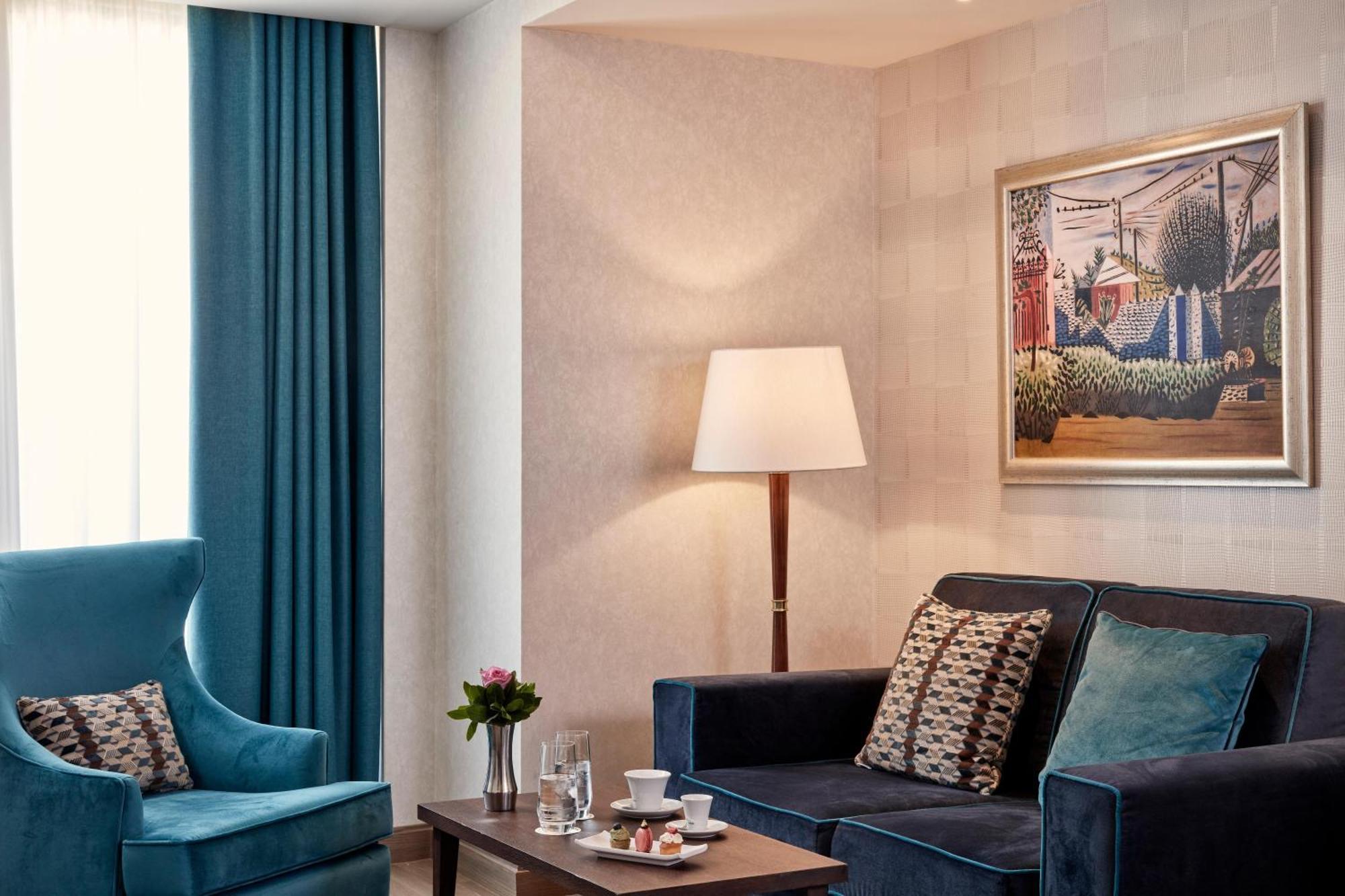 Electra Metropolis Hotel Athena Bagian luar foto The photo shows a stylish and modern living space. There is a dark blue couch adorned with decorative cushions, paired with a comfortable blue armchair. A coffee table in the center features a small floral arrangement and a couple of glassware items,