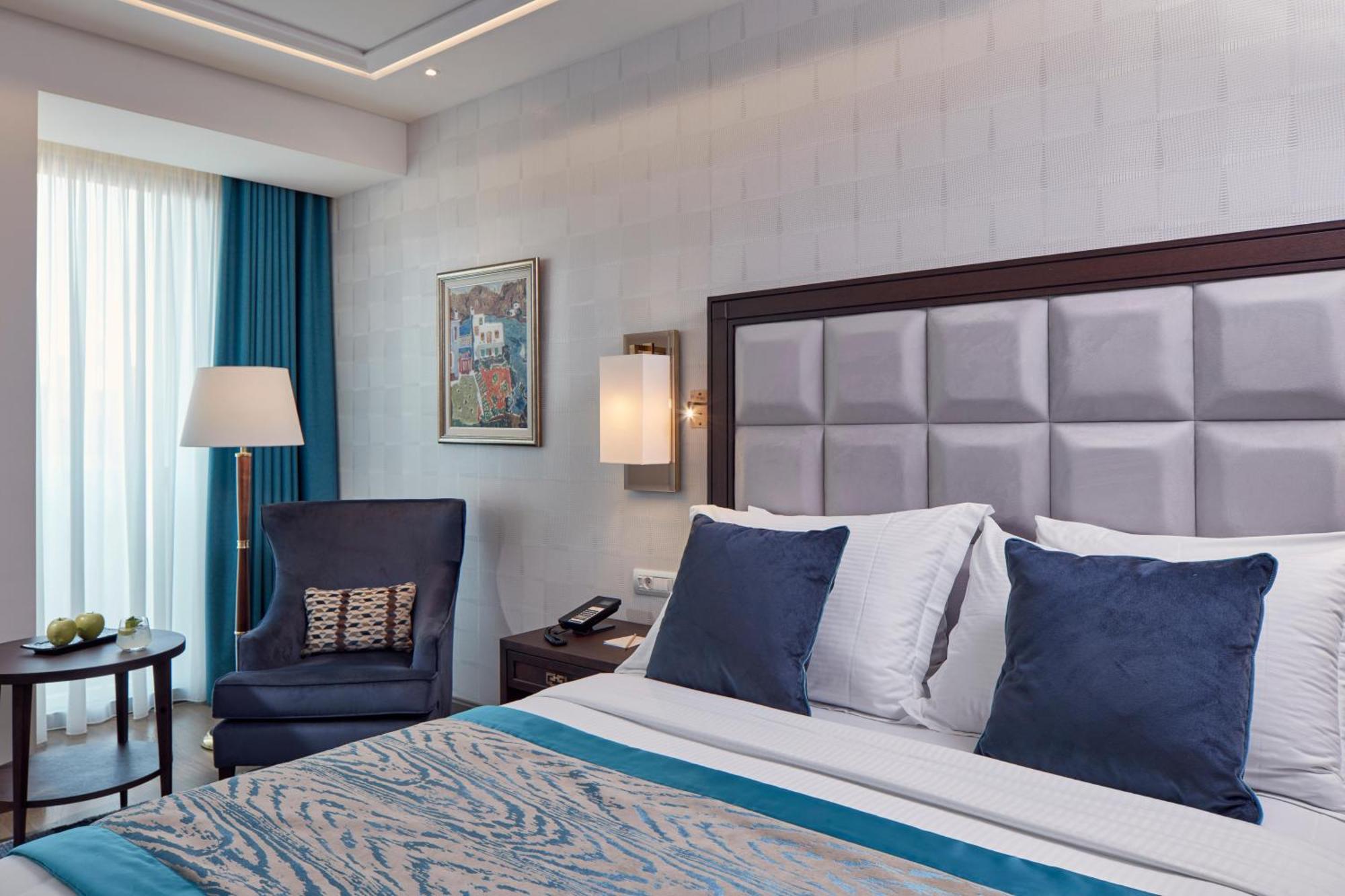 Electra Metropolis Hotel Athena Bagian luar foto The photo depicts a modern hotel room featuring a neatly made bed with white linens and blue accent pillows. There's a plush blue armchair next to a small table, which may hold some decorative items, including a bowl of green apples. A large window a