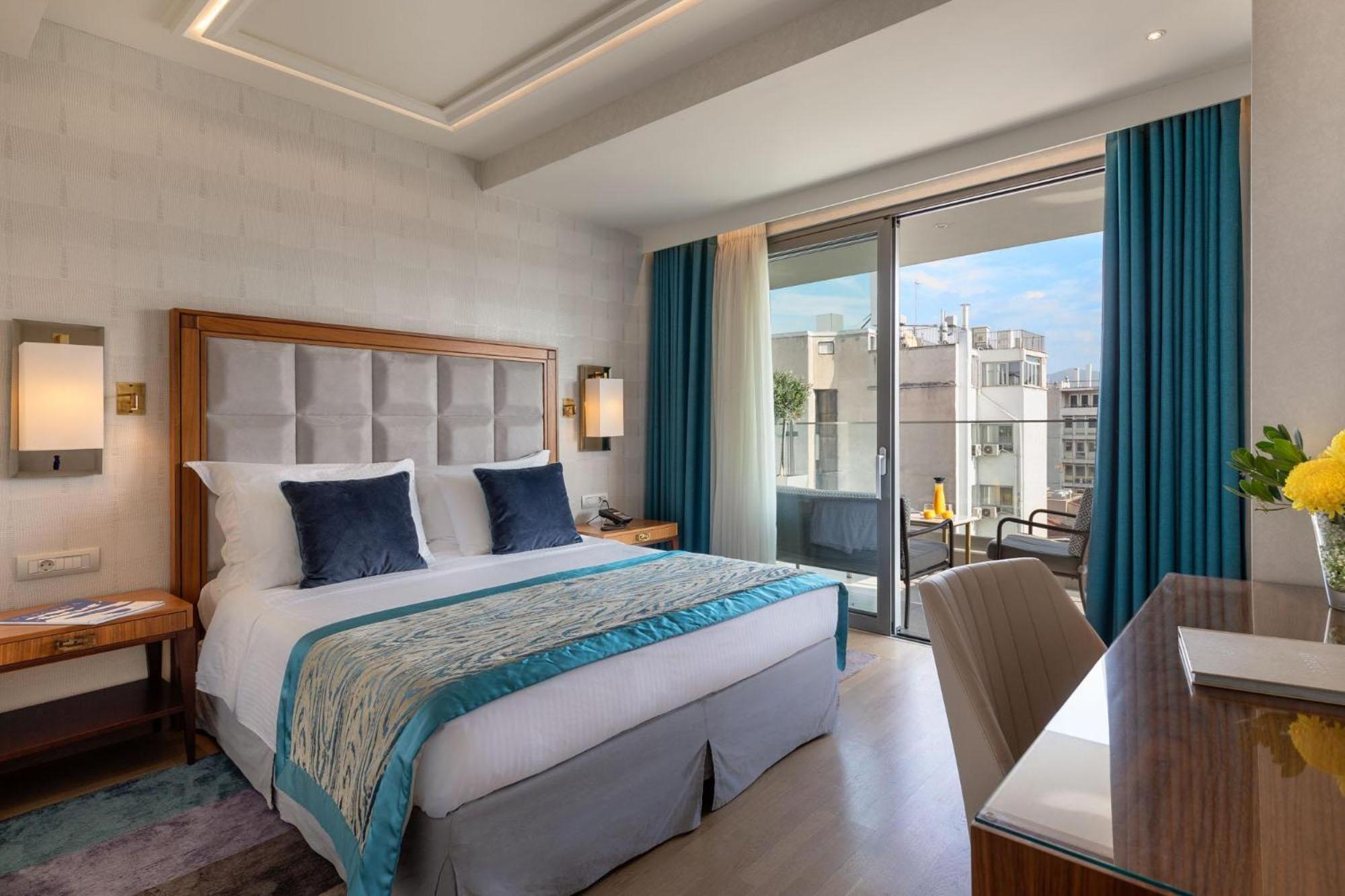 Electra Metropolis Hotel Athena Bagian luar foto The photo shows a modern hotel room featuring a queen-sized bed with a light-colored headboard. The bed is adorned with decorative pillows and a stylish throw blanket. There are two bedside lamps on either side of the bed. A set of floor-to-ceiling w