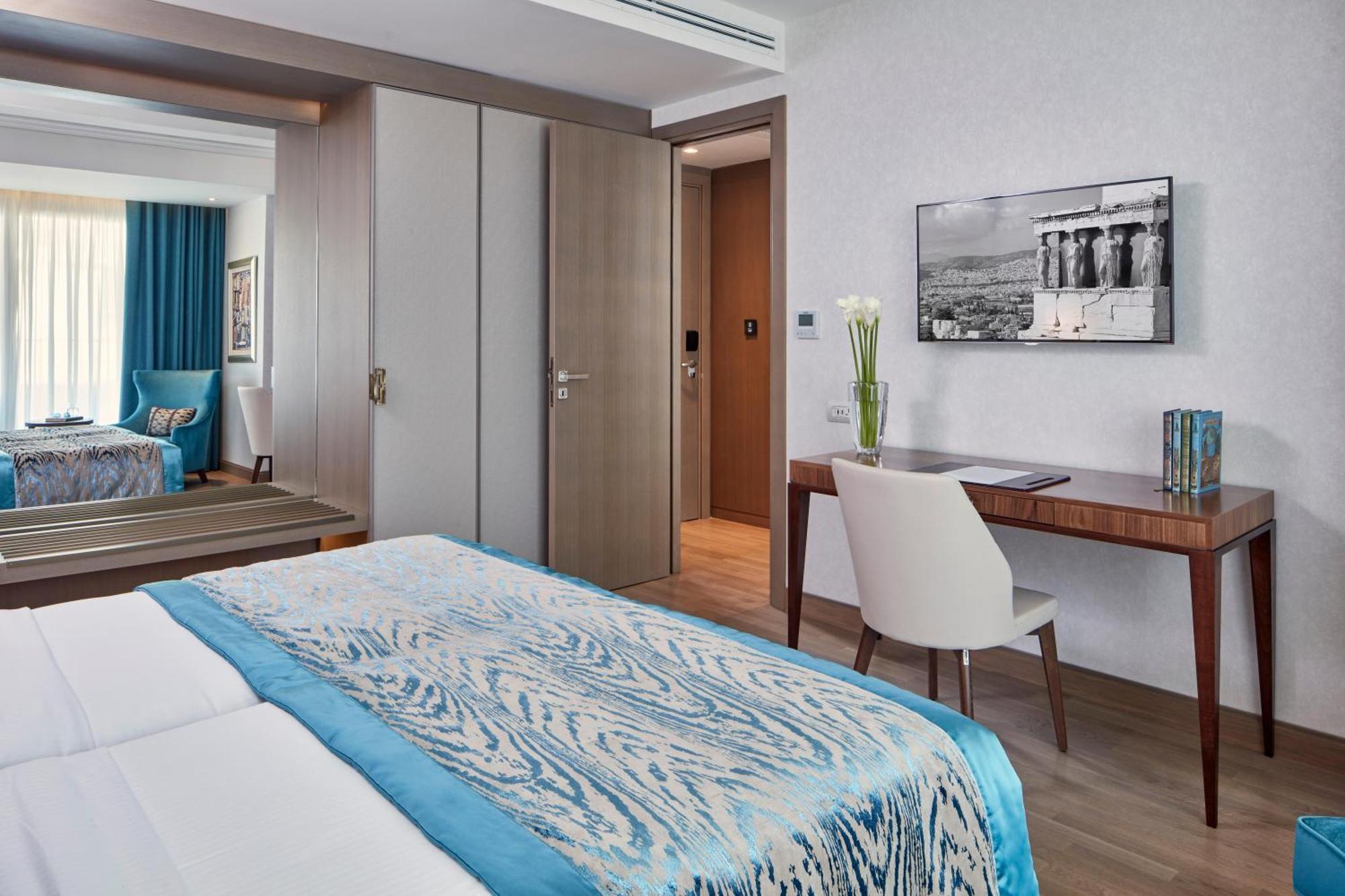 Electra Metropolis Hotel Athena Bagian luar foto The photo shows a modern hotel room with two beds covered in light-colored linens that have a blue patterned throw or blanket. There's a wooden desk and a white chair situated next to the wall, with a vase of flowers on the desk and a framed black-an