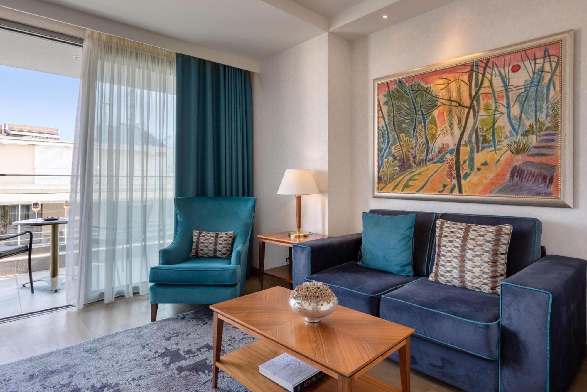 Electra Metropolis Hotel Athena Bagian luar foto The photo shows a modern living room area. There is a blue sofa with decorative pillows and a wooden coffee table in front of it. A light blue armchair is positioned next to the sofa. A painting with vibrant colors depicting a natural scene is hangin
