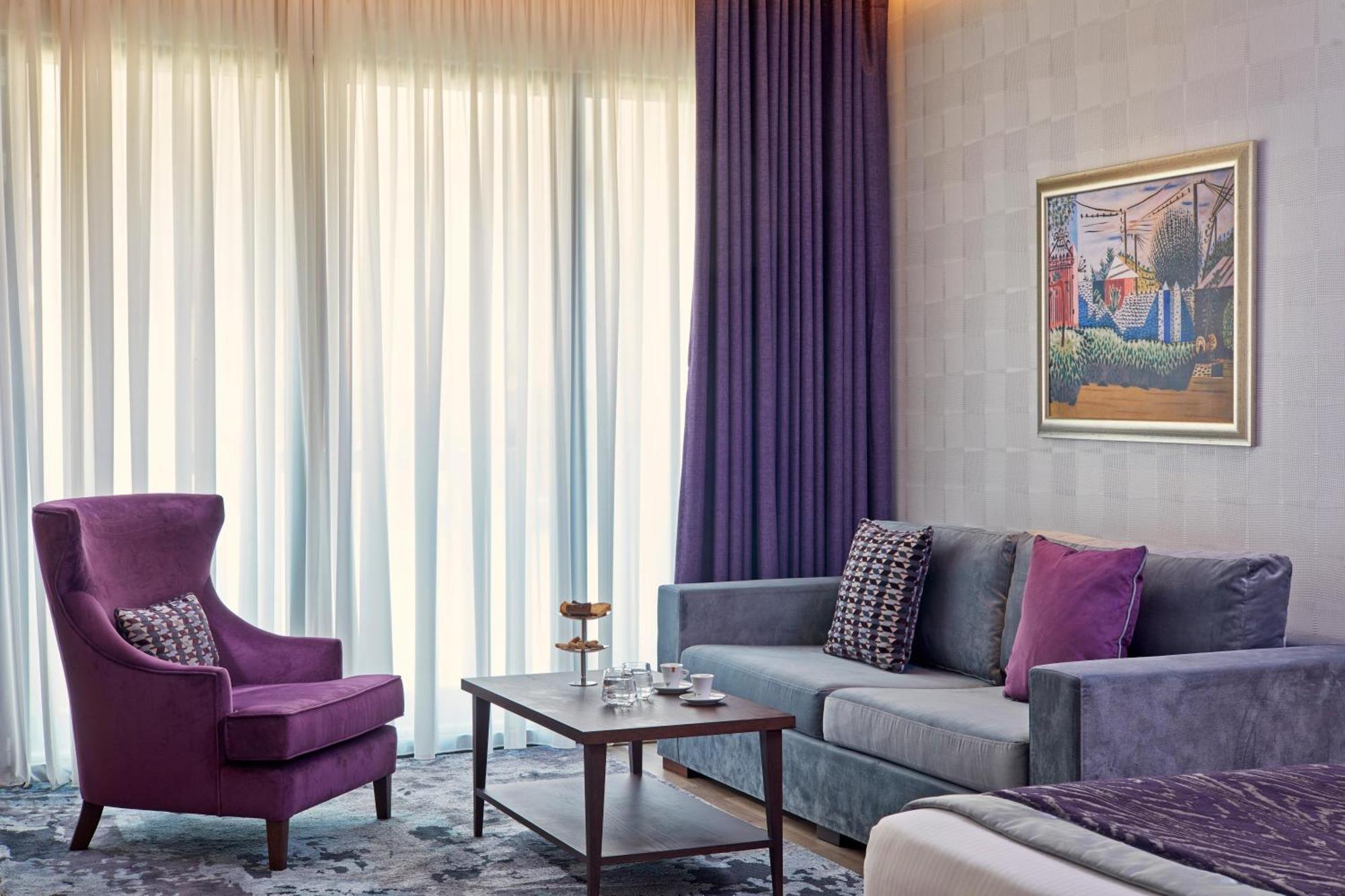 Electra Metropolis Hotel Athena Bagian luar foto The photo shows a stylish and modern hotel room or living area. It features large windows covered with sheer curtains that allow natural light to filter in, creating a bright atmosphere. 

On the left, there is a purple armchair with a contemporary d