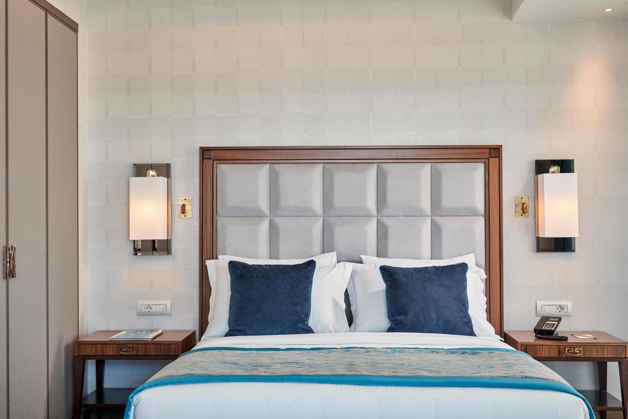 Electra Metropolis Hotel Athena Bagian luar foto The photo shows a well-designed hotel room with a cozy bed as the focal point. The bed has a soft, padded headboard in a light color, paired with four decorative pillows—two in a dark blue shade and two in white. On either side of the bed, there are 