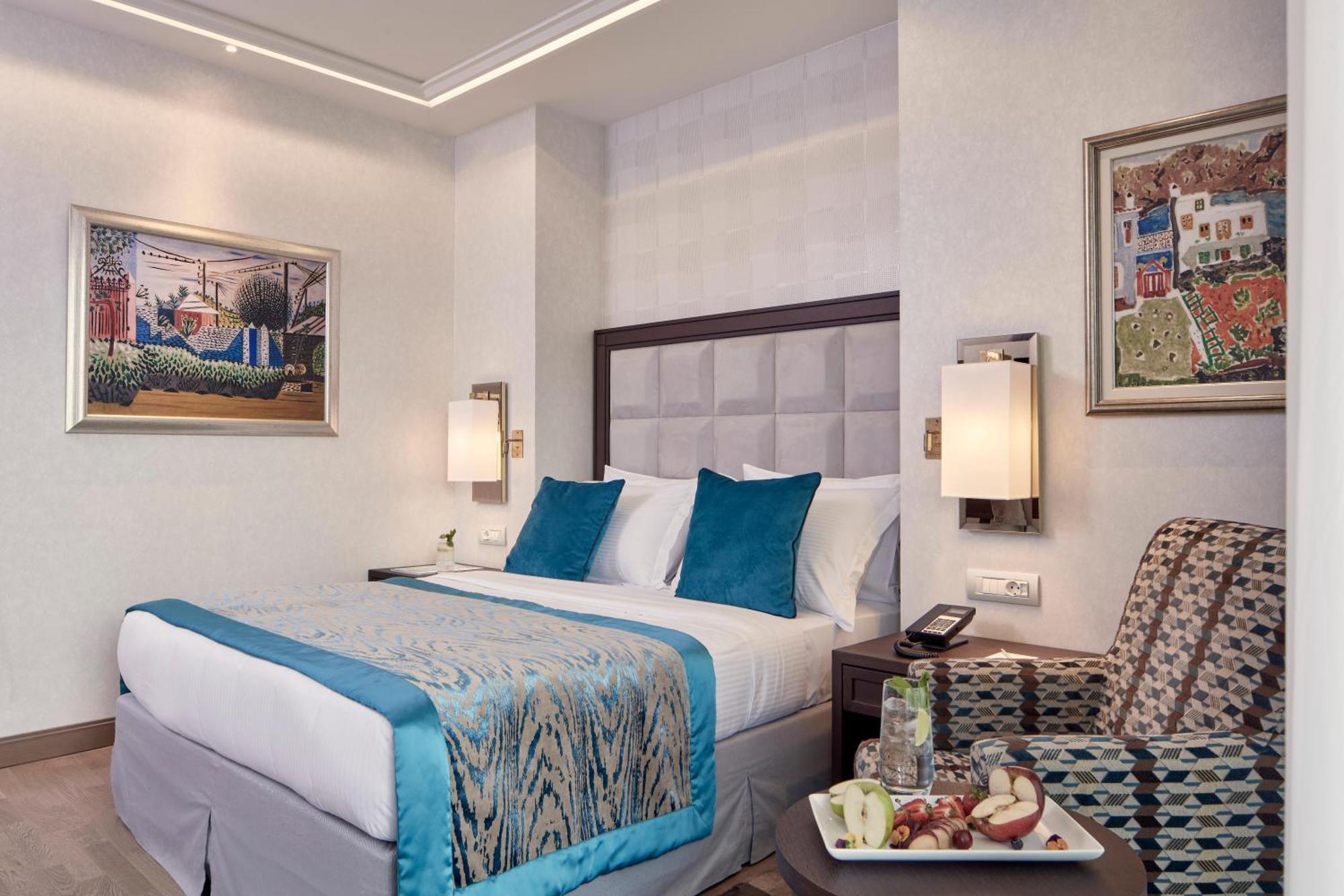Electra Metropolis Hotel Athena Bagian luar foto The photo shows a hotel room featuring a queen-sized bed with a light blue and patterned bedspread. The bed has white pillows with two accent pillows in turquoise. Beside the bed, there are contemporary lamps mounted on the wall. 

To the side, there
