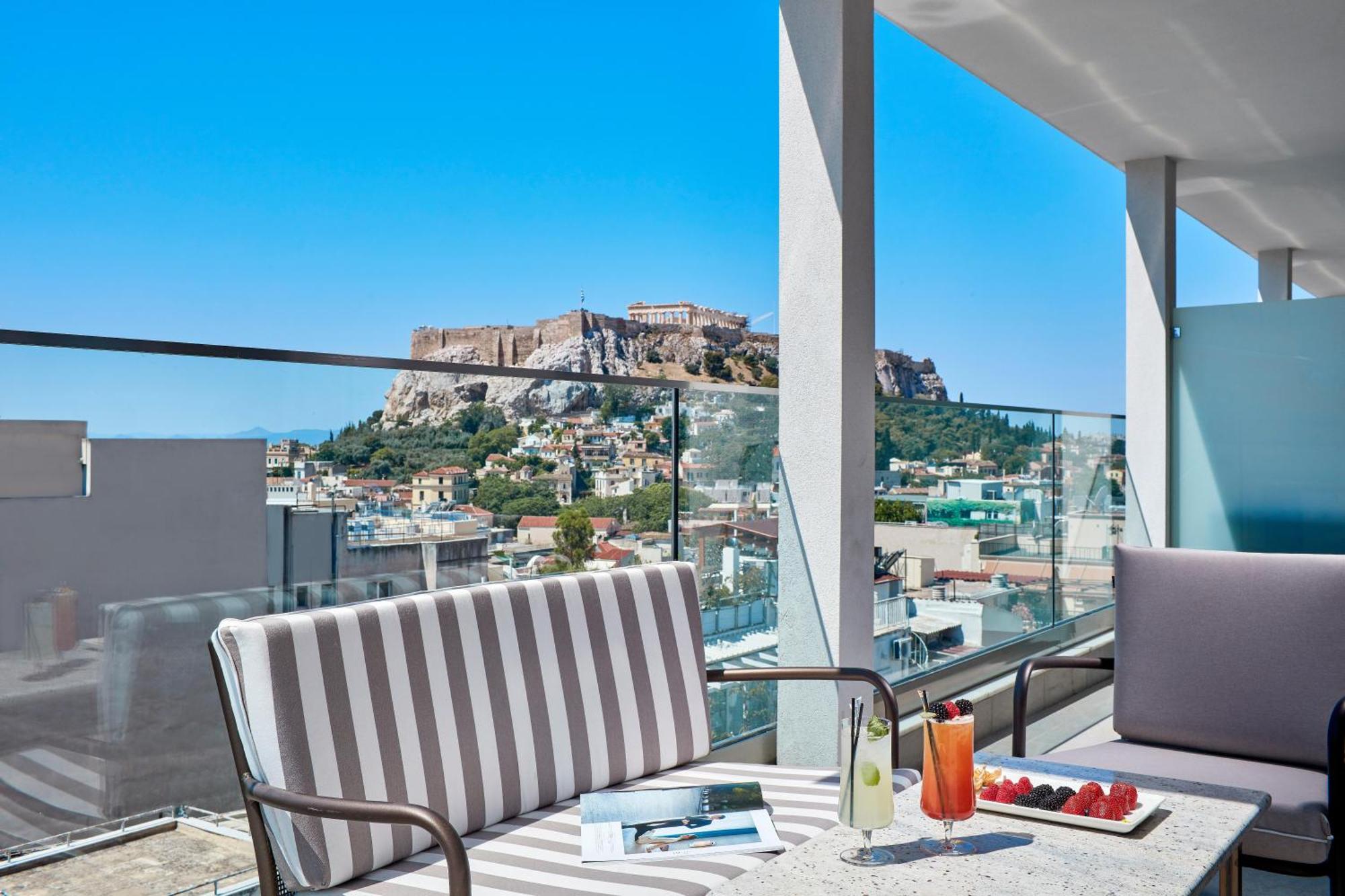 Electra Metropolis Hotel Athena Bagian luar foto The photo features a modern terrace or balcony with stylish seating arrangements. There are two cushioned chairs, one with striped fabric. A table in front holds a beverage, possibly a cocktail, and some fresh strawberries. The backdrop showcases a b