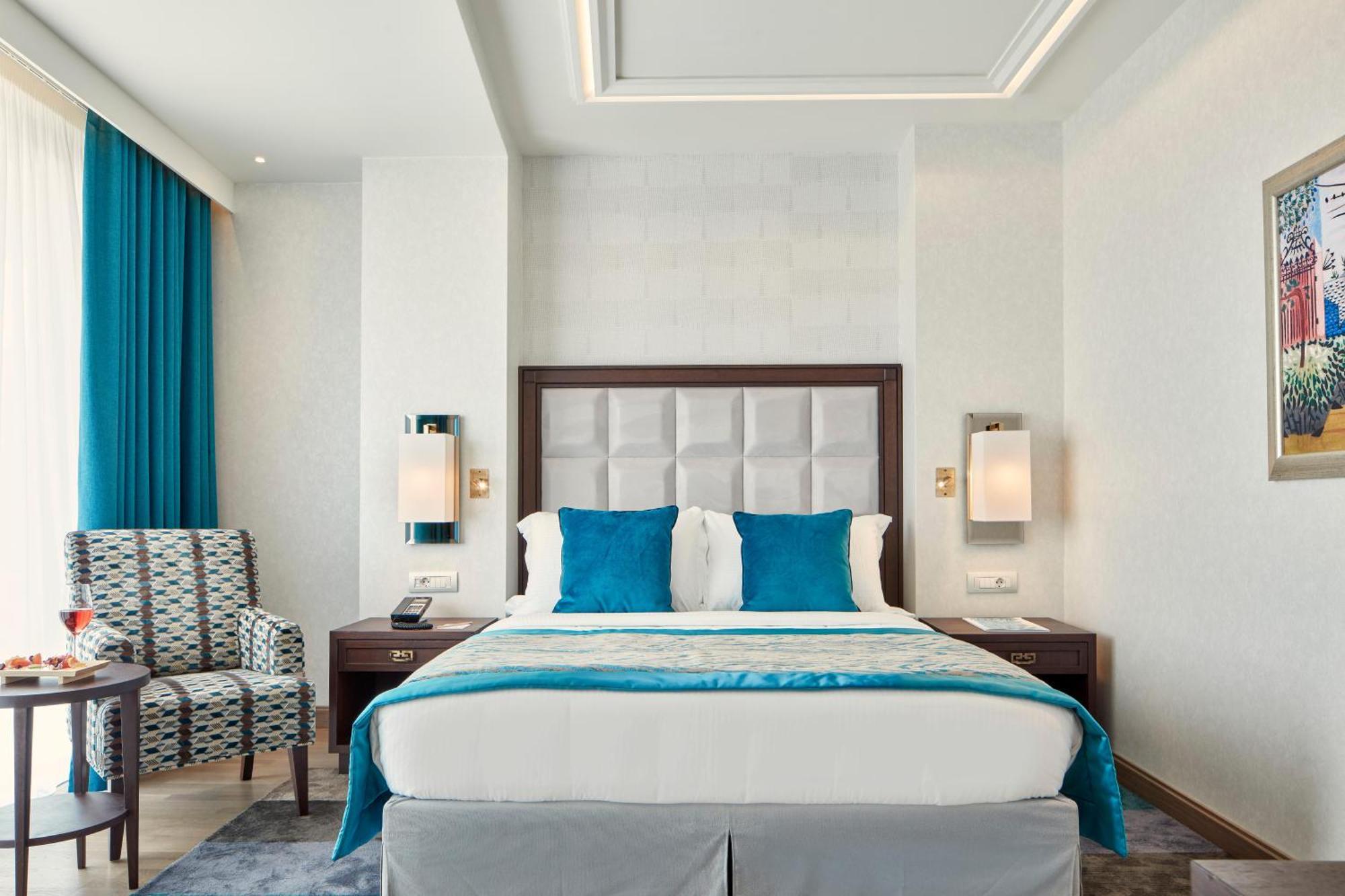 Electra Metropolis Hotel Athena Bagian luar foto The photo shows a modern hotel room. In the center, there is a large bed with a white and light blue bedding set. The headboard is upholstered in a light gray material. On either side of the bed, there are wall-mounted lamps. There is a stylish chair