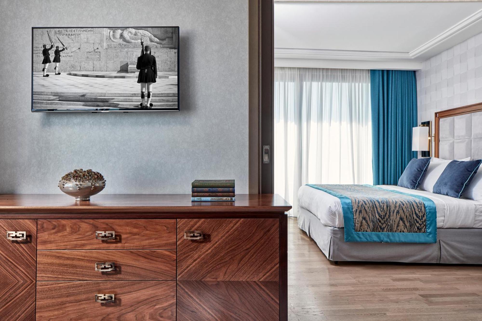 Electra Metropolis Hotel Athena Bagian luar foto The photo shows a modern, stylish hotel room or suite. In the foreground, there is a wooden dresser with several drawers, and on top, there is a decorative piece, a few books, and a television mounted on the wall displaying a black-and-white image of