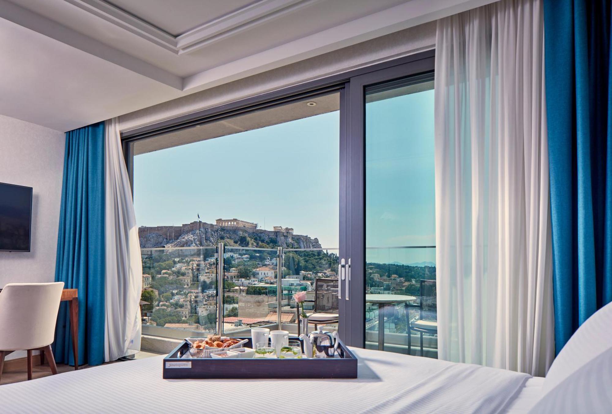 Electra Metropolis Hotel Athena Bagian luar foto The photo shows a modern hotel room with a large window offering a view of a historical site, likely a fortress or ancient ruins, situated on a hill. The interior features a neatly made bed with a tray placed on it, displaying various items that coul