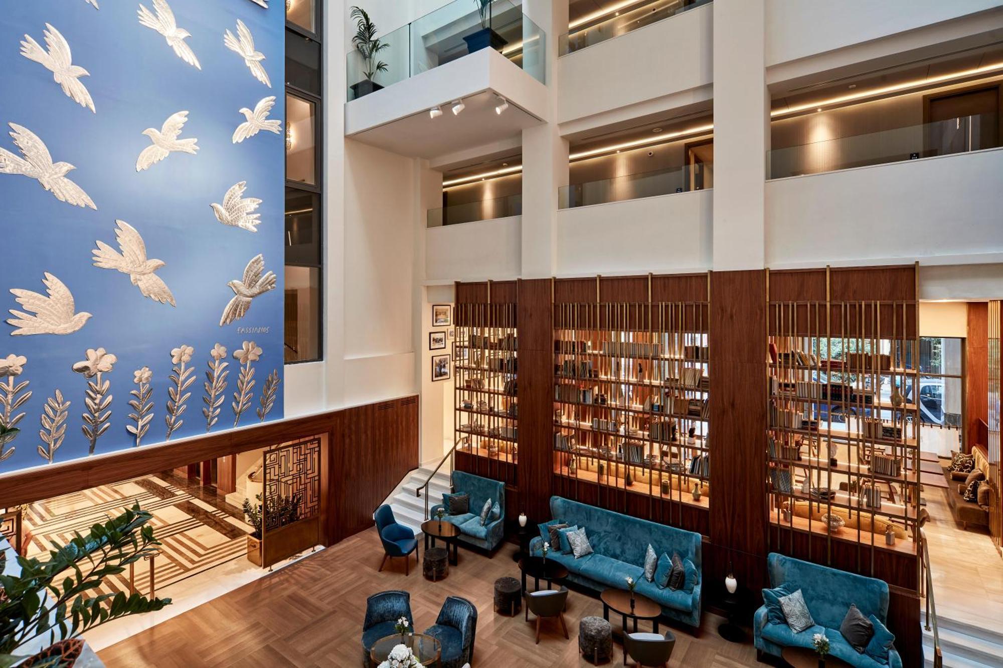 Electra Metropolis Hotel Athena Bagian luar foto The photo depicts a modern, stylish interior space, likely a hotel or a lobby area. The design features a high ceiling with ample natural light. 

To the left, there is a large blue wall adorned with decorative white birds, adding a vibrant touch to 