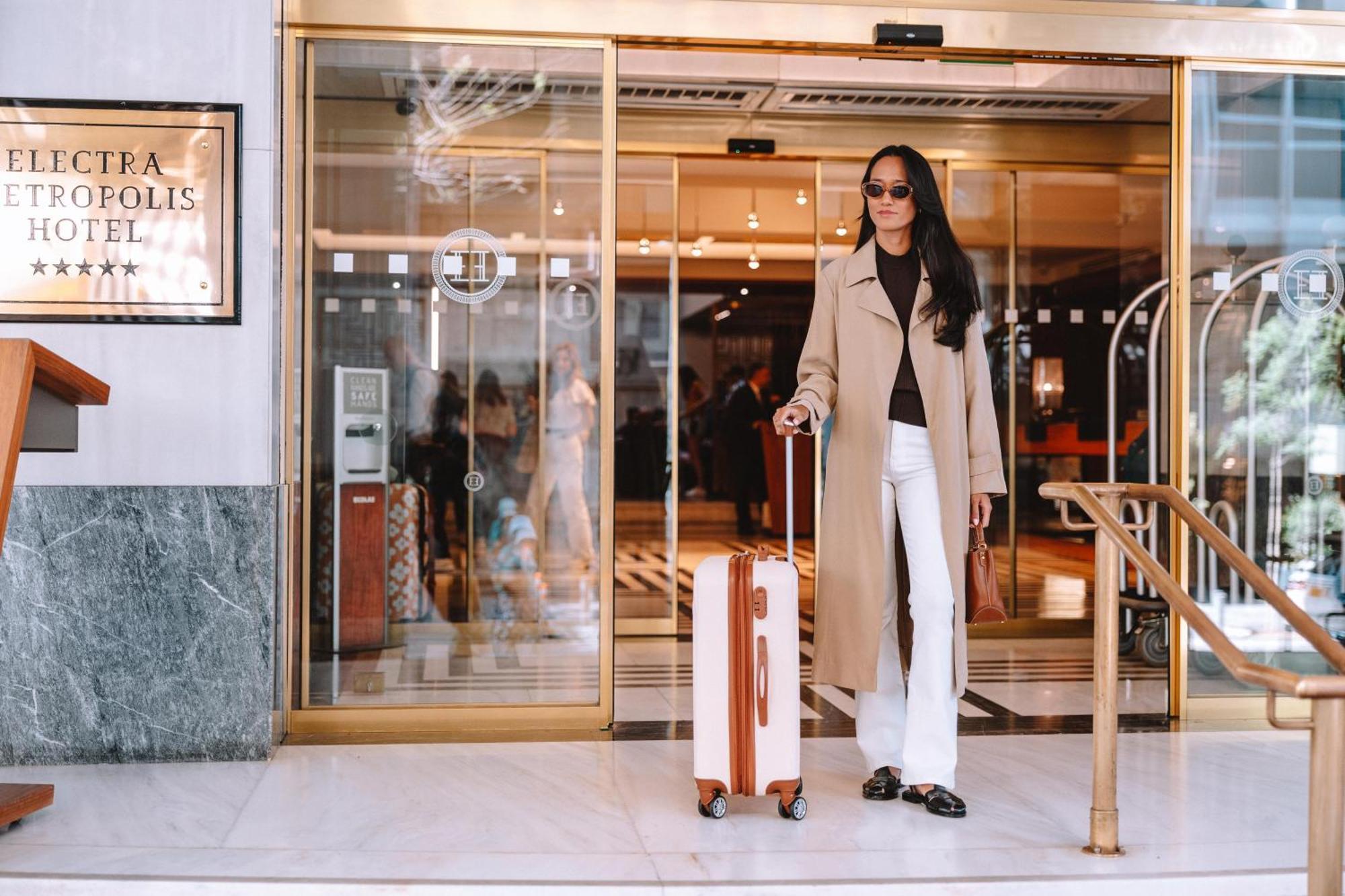 Electra Metropolis Hotel Athena Bagian luar foto The photo shows a fashionable individual standing outside a hotel entrance. The person is wearing a long beige coat over a dark top, paired with white pants. They are holding a stylish suitcase with a wooden design. The hotel has large glass doors, a