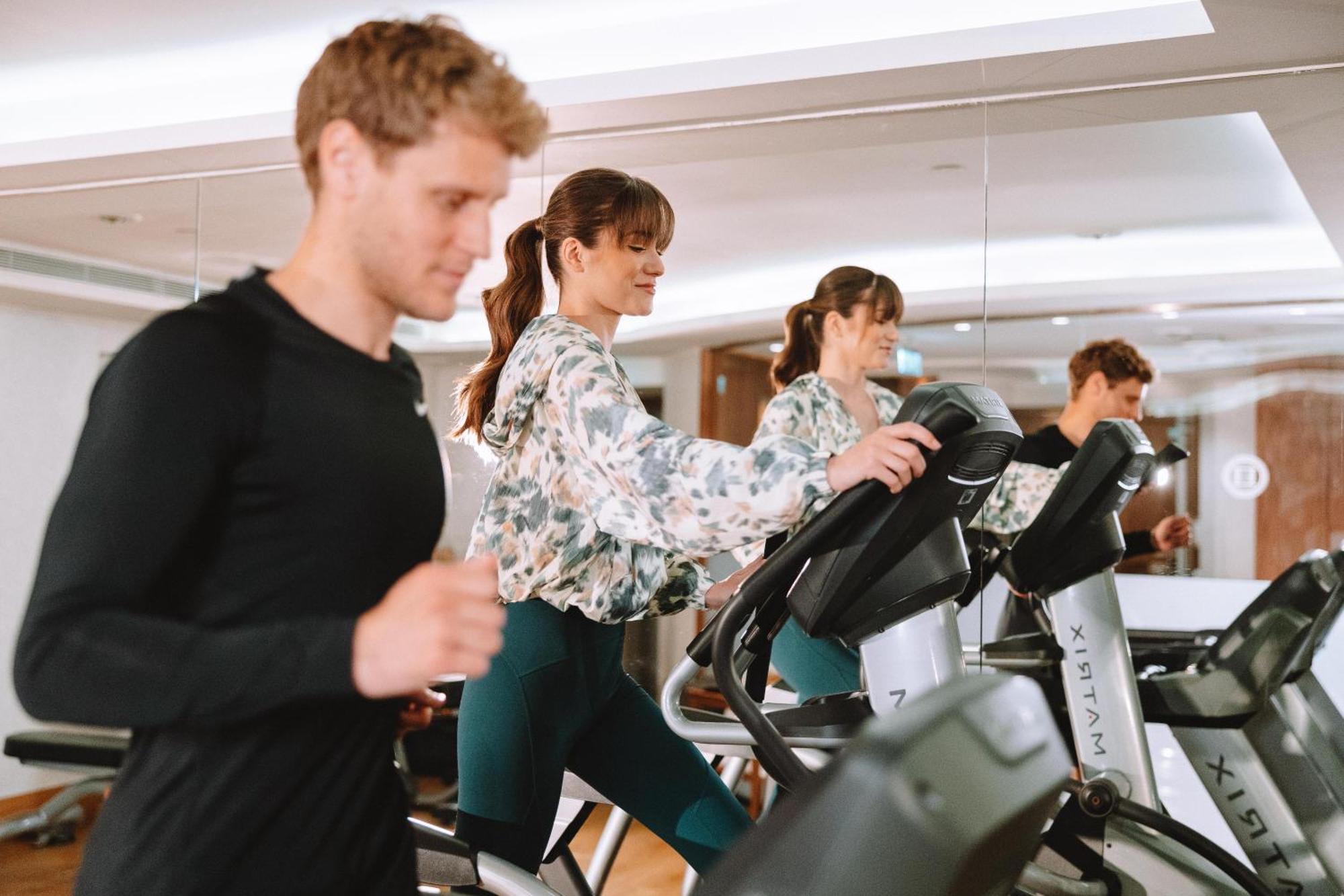Electra Metropolis Hotel Athena Bagian luar foto The photo shows two people exercising in a gym. One man is running on a treadmill, while a woman is using an elliptical machine next to him. The woman is wearing a floral blouse and leggings, and the man is dressed in a black athletic shirt. They bot