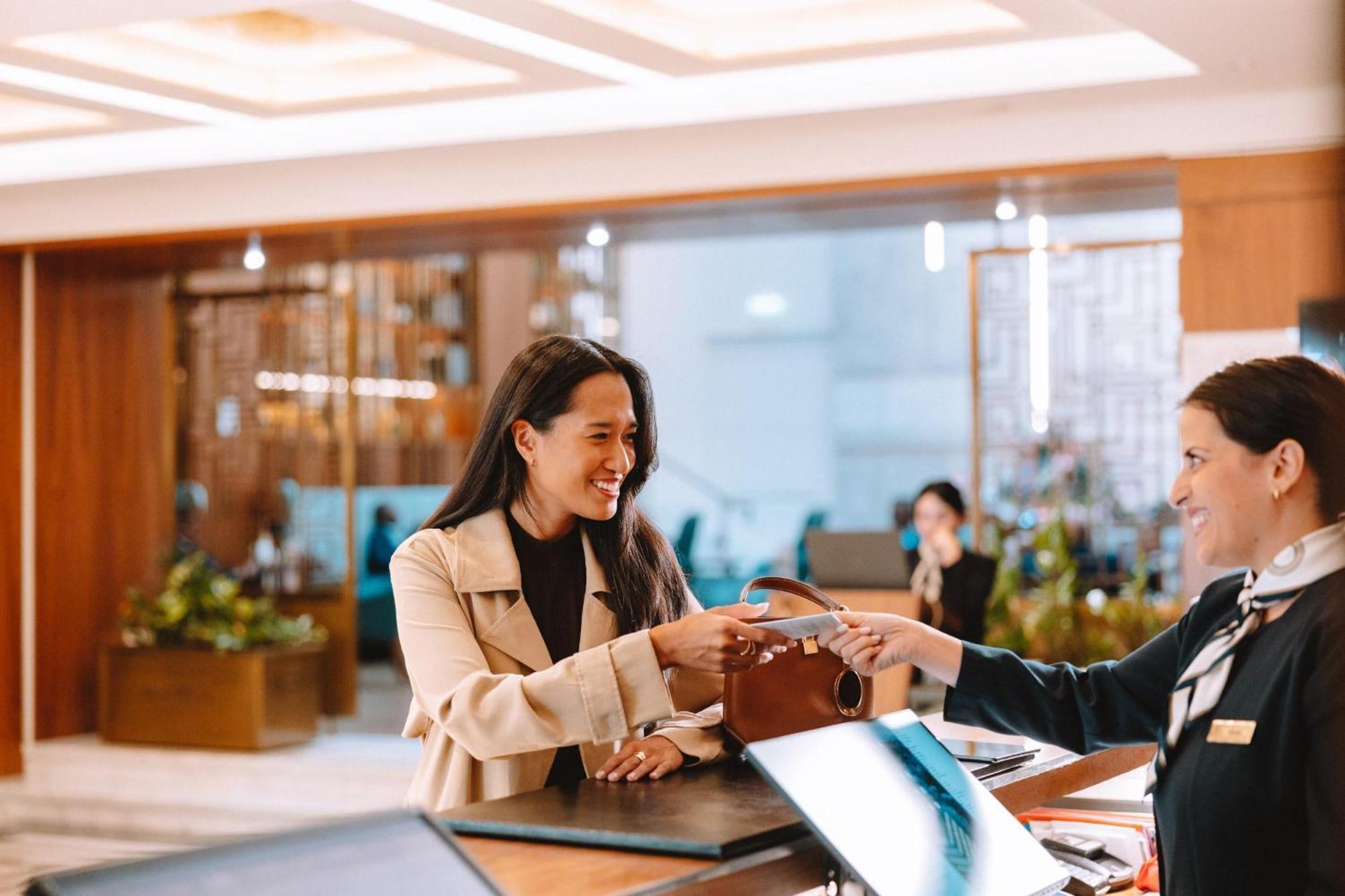 Electra Metropolis Hotel Athena Bagian luar foto The photo shows a scene at a reception area, likely in a hotel. A woman wearing a beige coat is smiling as she hands over a brown bag to a receptionist. The receptionist, dressed in a formal uniform, is receiving the bag with a friendly expression. I